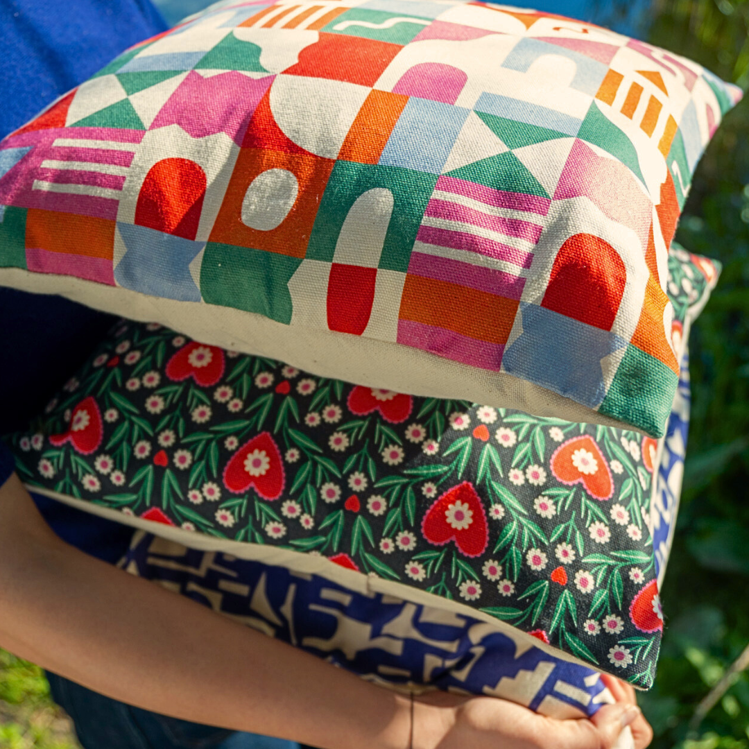 Personne tenant trois coussins carrés design 40x40, aux motifs illustrés colorés par Claire Iglesias, en extérieur au bord de l’eau – collection Sézons.