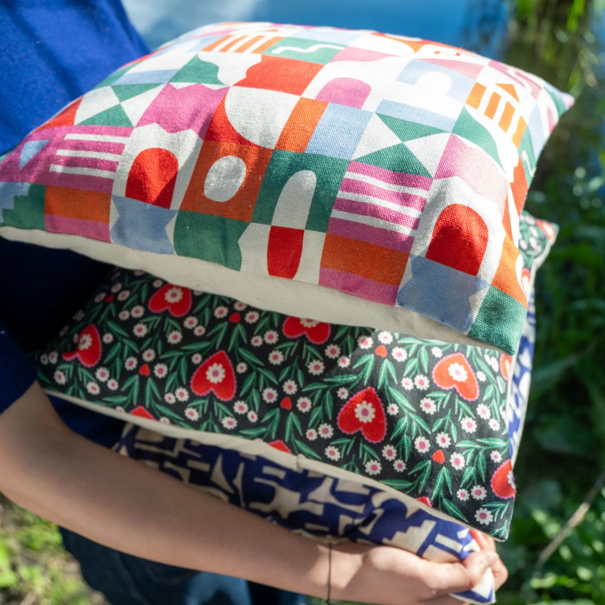 Personne tenant trois coussins carrés design 40x40, aux motifs illustrés colorés par Claire Iglesias, en extérieur au bord de l’eau – collection Sézons.